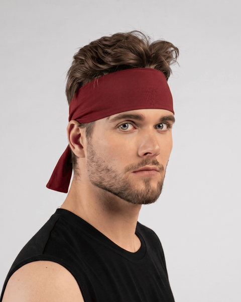 A fair-skinned man with brown hair and a short beard is wearing a black sleeveless shirt and a deep red tie headband. He is posed in a three-quarter profile while looking directly at the camera, with the tied tail of the headband hanging down behind his head against a light gray background.