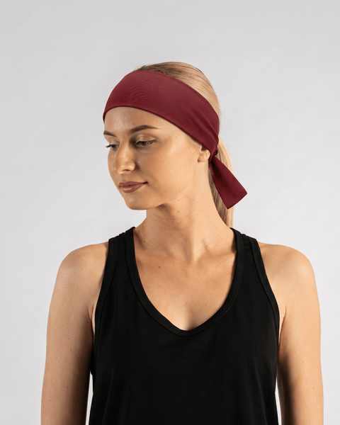 A fair-skinned woman with blonde hair pulled back into a ponytail wearing a black tank top and a deep red (royal cherry) tie headband. She is looking down and to the side, with the tied tails of the headband clearly visible at the nape of her neck against a light gray background.
