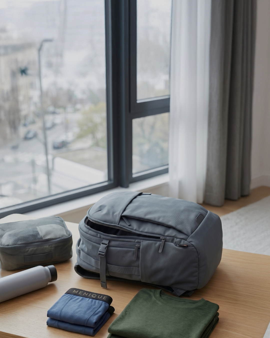 A lifestyle shot showing travel preparation. On a light wooden bench, a grey backpack sits partially open next to a grey packing cube and a grey water bottle. Neatly folded in front of the bag are a pair of folded blue Menique boxer briefs and a folded dark green t-shirt. The background shows a portion of a room with a window, curtains, and light wood flooring.