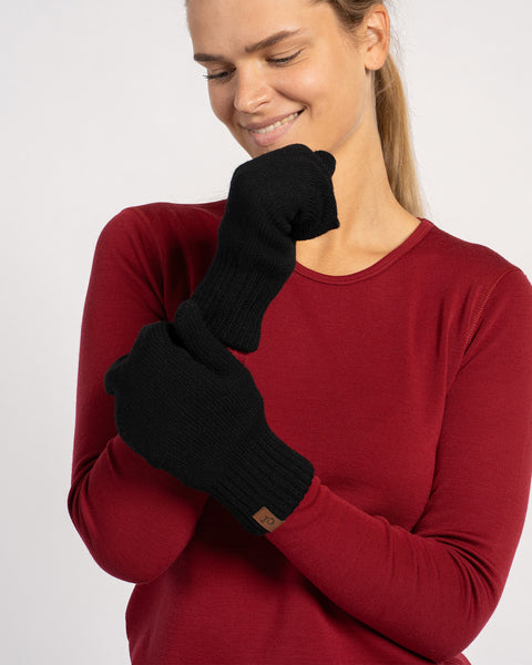 Woman wearing black knitted merino wool mittens with a red long-sleeve top, smiling and crossing arms.