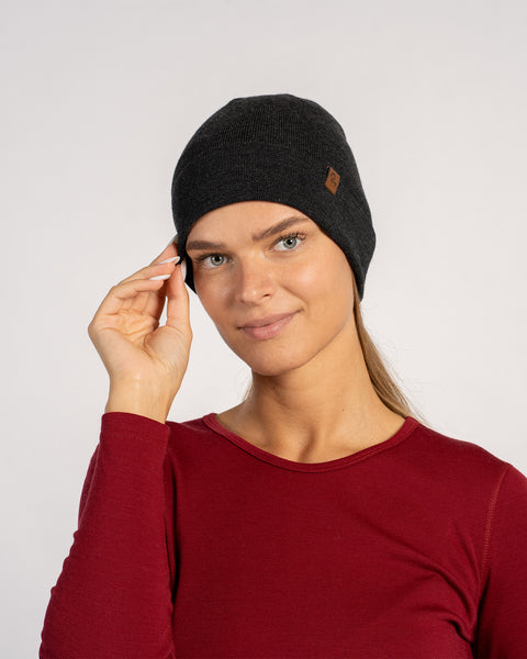 Woman in red shirt adjusting a dark gray knitted Merino wool beanie, front view.