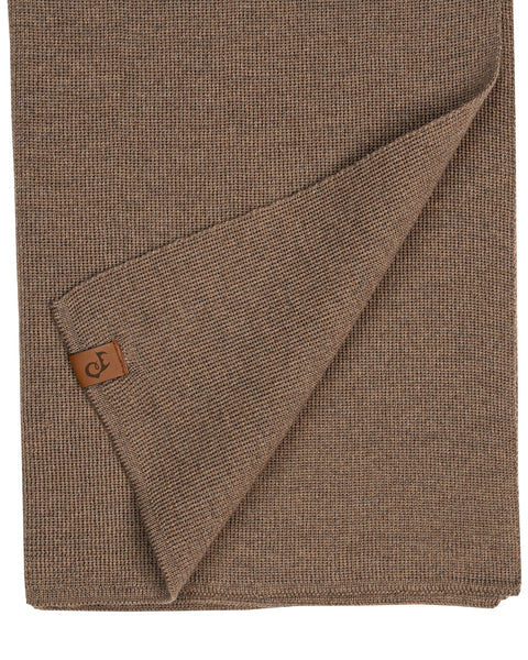 A close-up flat lay of a light brown or taupe knitted merino wool scarf. The bottom edge is folded up diagonally, displaying a small, rectangular brown leather logo patch attached near the hem. The knit pattern is clearly detailed. The background is pure white.