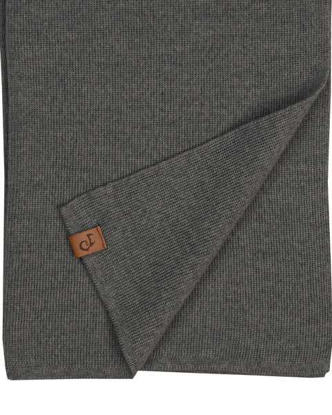 A close-up flat lay of a dark gray knitted merino wool scarf. The bottom edge is folded up diagonally to reveal a small, rectangular brown leather logo patch sewn near the hem. The fine knit texture is visible. The background is pure white.