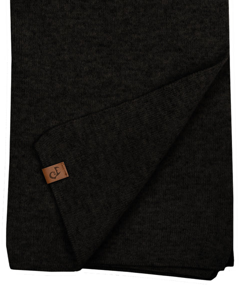 A close-up flat lay of a black knitted scarf. A corner of the hem is folded up diagonally, displaying a small, square brown leather logo patch. The texture of the fine knit is visible against the pure white background.
