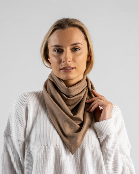 A studio portrait of a woman looking directly at the camera. She is wearing a large, light beige bandana draped around her neck like a cowl, with her left hand resting lightly on the fabric near her collarbone. She is wearing a textured white sweater, and the background is a solid light grey.