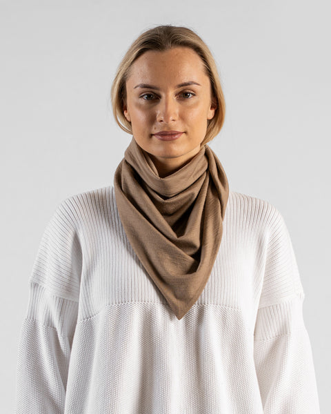 A studio portrait of a woman looking directly at the camera with a subtle smile. She is wearing a large, light beige bandana draped around her neck like a cowl, with the triangle point hanging down in front. She is wearing a textured white sweater against a plain light grey background.