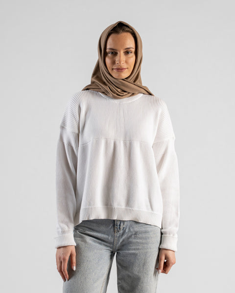 A fair-skinned woman wearing a white textured sweater and light blue jeans, with a large beige bandana draped over her head and wrapped around her neck like a headscarf. She is standing and looking directly at the camera against a plain light gray background.