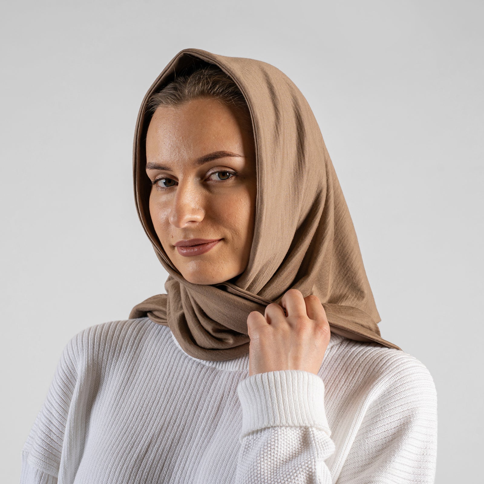 A studio portrait of a woman looking off to the left side of the frame. She is wearing a small, light beige bandana tied loosely around her neck like a scarf. She is wearing a textured white sweater, and the background is a solid light grey.