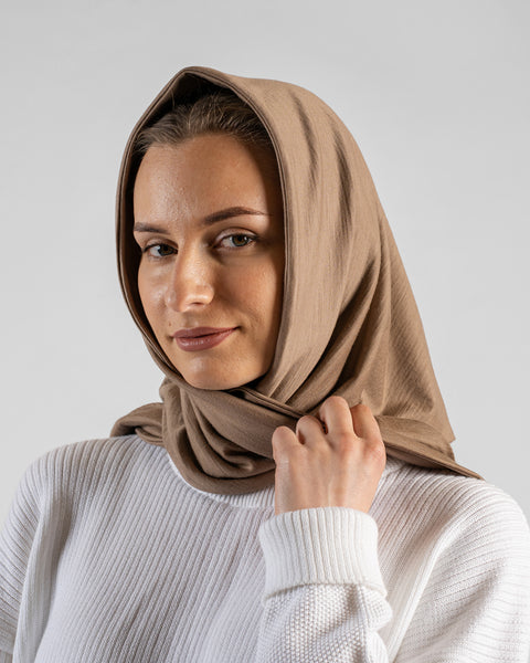 A close-up studio portrait of a woman looking directly at the camera with a soft smile against a plain light grey background. She is wearing a white textured sweater and a large beige bandana draped loosely over her head like a headscarf, holding the fabric gently near her collar.