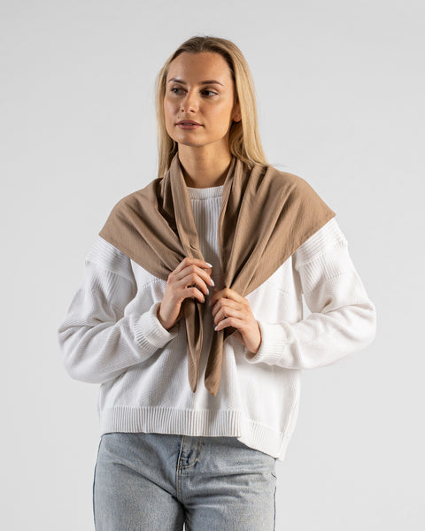 A studio portrait of a blonde woman looking off to the left against a plain light grey background. She is wearing a white textured sweater, light blue jeans, and a large beige bandana draped over her shoulders, gently holding the two front ends with her hands.