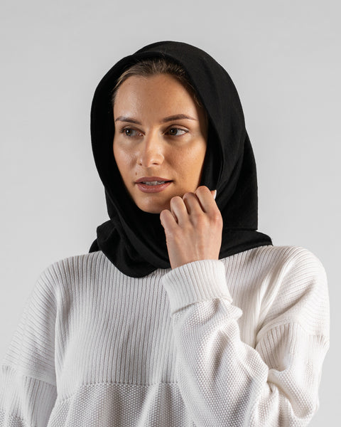 A studio portrait of a woman looking off to the left side of the frame. She is wearing a large, solid black bandana draped over her head like a hood. Her right hand is gently touching the edge of the fabric near her chin. She is wearing a textured white sweater, and the background is a solid light grey.
