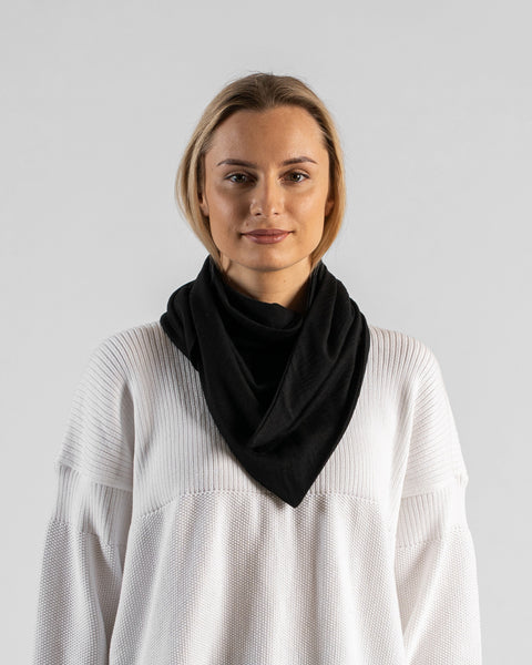 A studio portrait of a woman looking directly at the camera with a subtle smile. She is wearing a large, solid black bandana draped around her neck like a cowl, with the triangle point hanging down in front. She is wearing a textured white sweater against a plain light grey background.