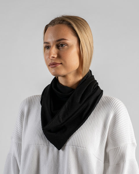 A fair-skinned woman with blonde hair pulled back, wearing a white textured sweater. She is wearing a large black bandana draped loosely around her neck, coming to a point in the front. She is looking off to the side against a plain light gray background.