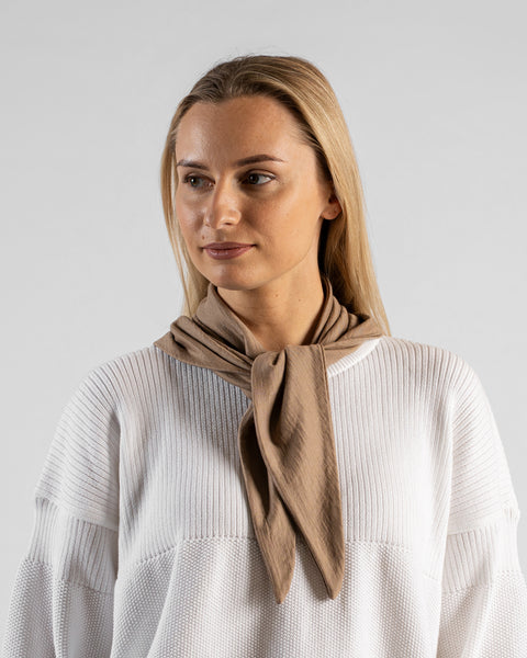 A studio portrait of a woman looking off to the left side of the frame. She is wearing a small, light beige bandana tied loosely around her neck like a scarf. She is wearing a textured white sweater, and the background is a solid light grey.