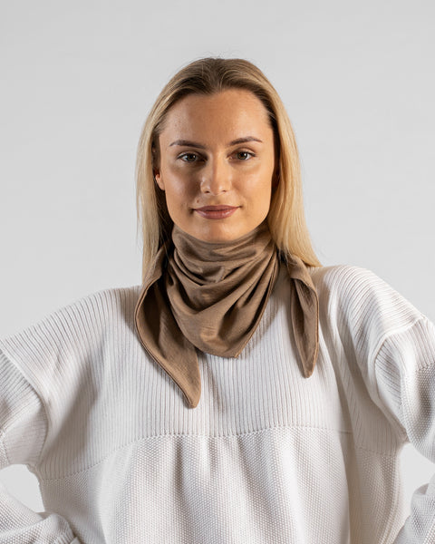 A studio portrait of a woman looking directly at the camera. She is wearing a small, light beige bandana tied around her neck with the triangle point hanging down in front. Her hands are resting on her hips. She is wearing a textured white sweater against a plain light grey background.