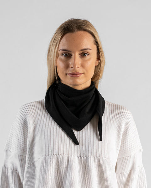 A studio portrait of a woman looking directly at the camera with a gentle expression. She is wearing a small, solid black bandana tied around her neck, with the triangle point and tied ends hanging down in front. She is wearing a textured white sweater against a plain light grey background.