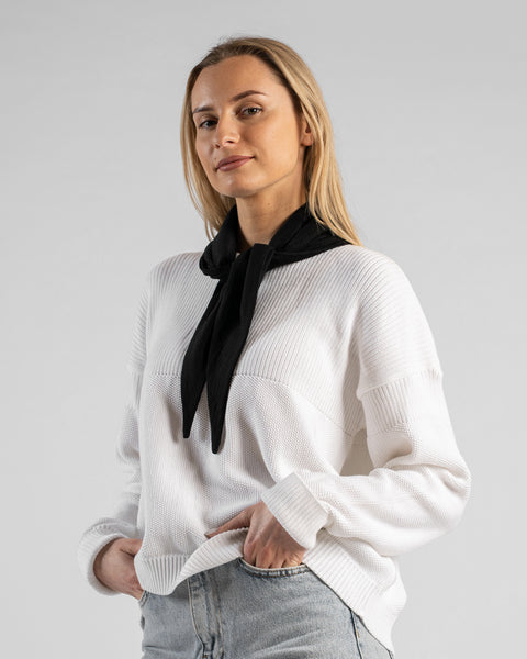 A studio portrait of a woman standing in a relaxed pose, looking off to the left side of the frame with one hand in the pocket of her light blue jeans. She is wearing a small, solid black bandana tied loosely around her neck with the ends hanging down. She is wearing a textured white sweater against a plain light grey background.