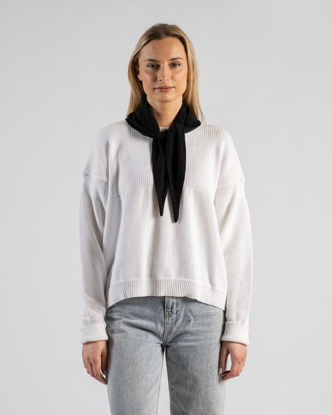 A fair-skinned woman with blonde hair wearing a white textured sweater and light blue jeans. She is wearing a small black bandana tied around her neck like a neckerchief, with the knot and two pointed ends hanging down in the front. She is looking directly at the camera against a plain light gray background.