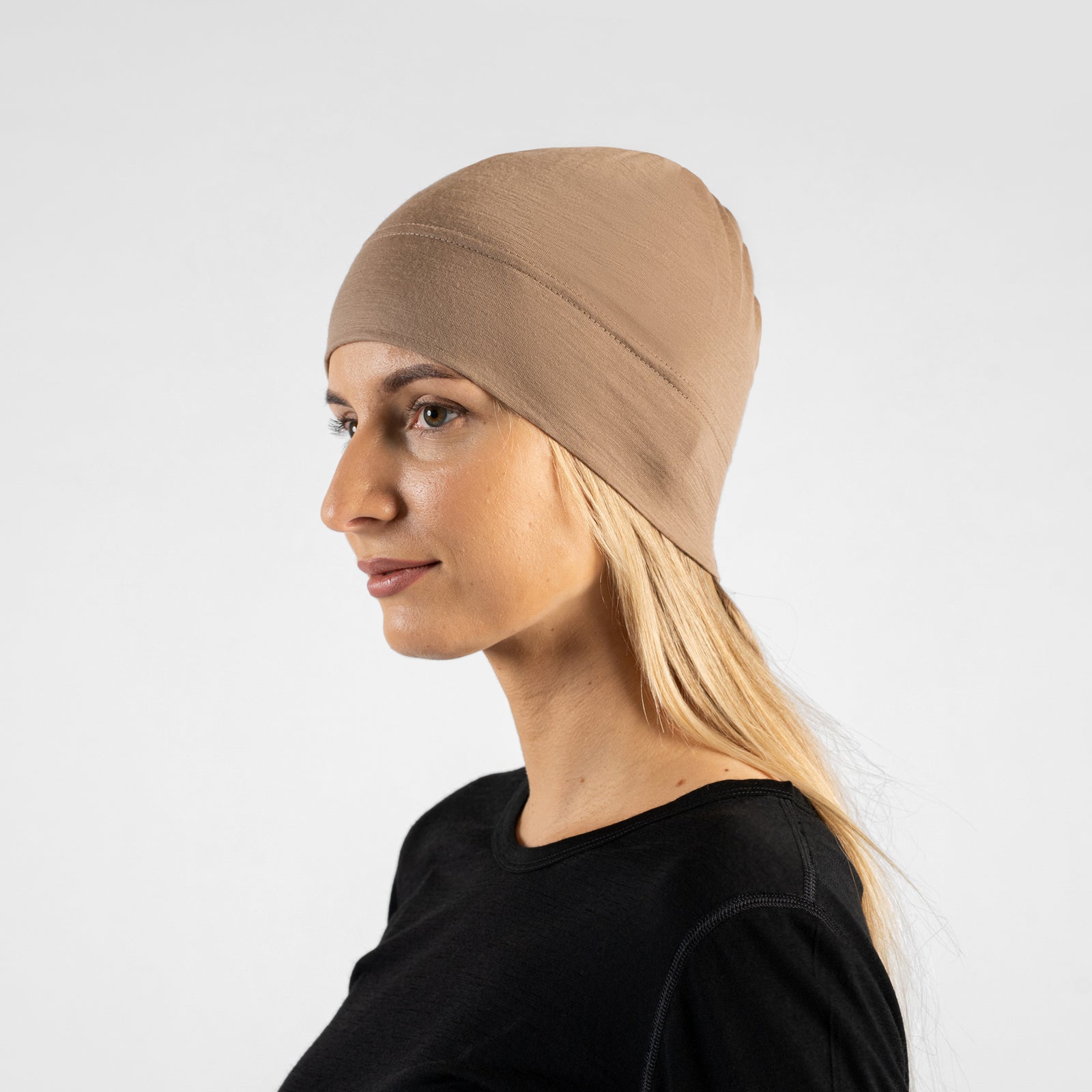 A studio portrait of a woman in a profile view, looking off to the left side of the frame. She is wearing a fitted, dark red (royal cherry) skull cap over her blonde hair and a black top. The background is a solid light grey.