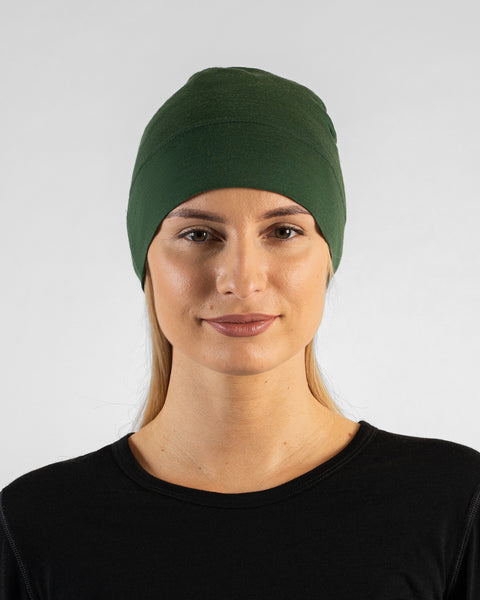 A studio portrait of a woman looking directly at the camera. She is wearing a fitted, dark green skull cap over her blonde hair and a black top. The background is a solid light grey.