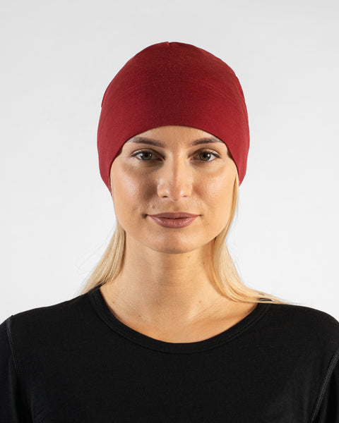 A studio portrait of a woman looking directly at the camera with a subtle smile. She is wearing a fitted, dark red (royal cherry) skull cap over her blonde hair and a black top. The background is a solid light grey.
