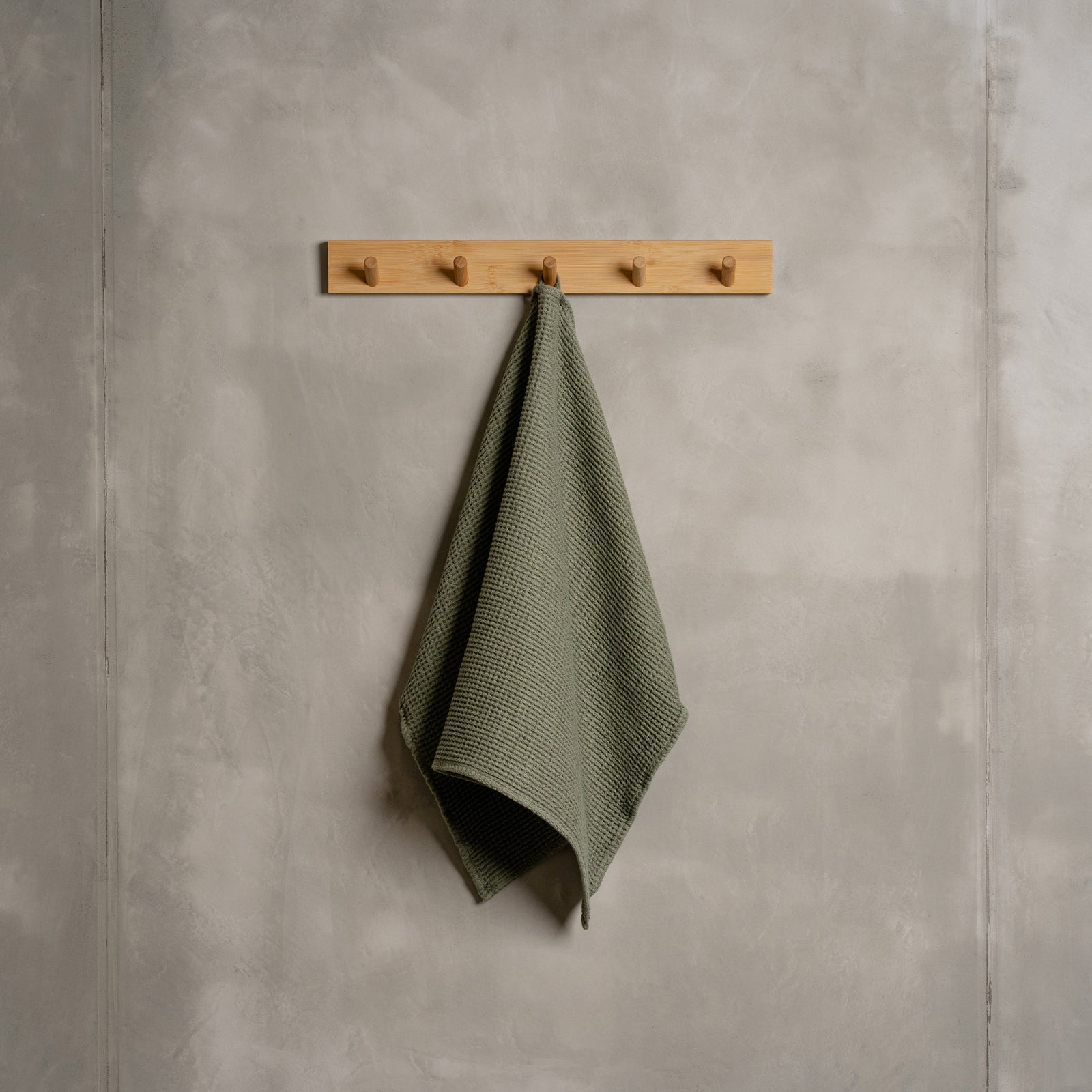 A gray wall with a wooden hanger. In the middle of it hangs a waffle hand washcloth in sand brown color.