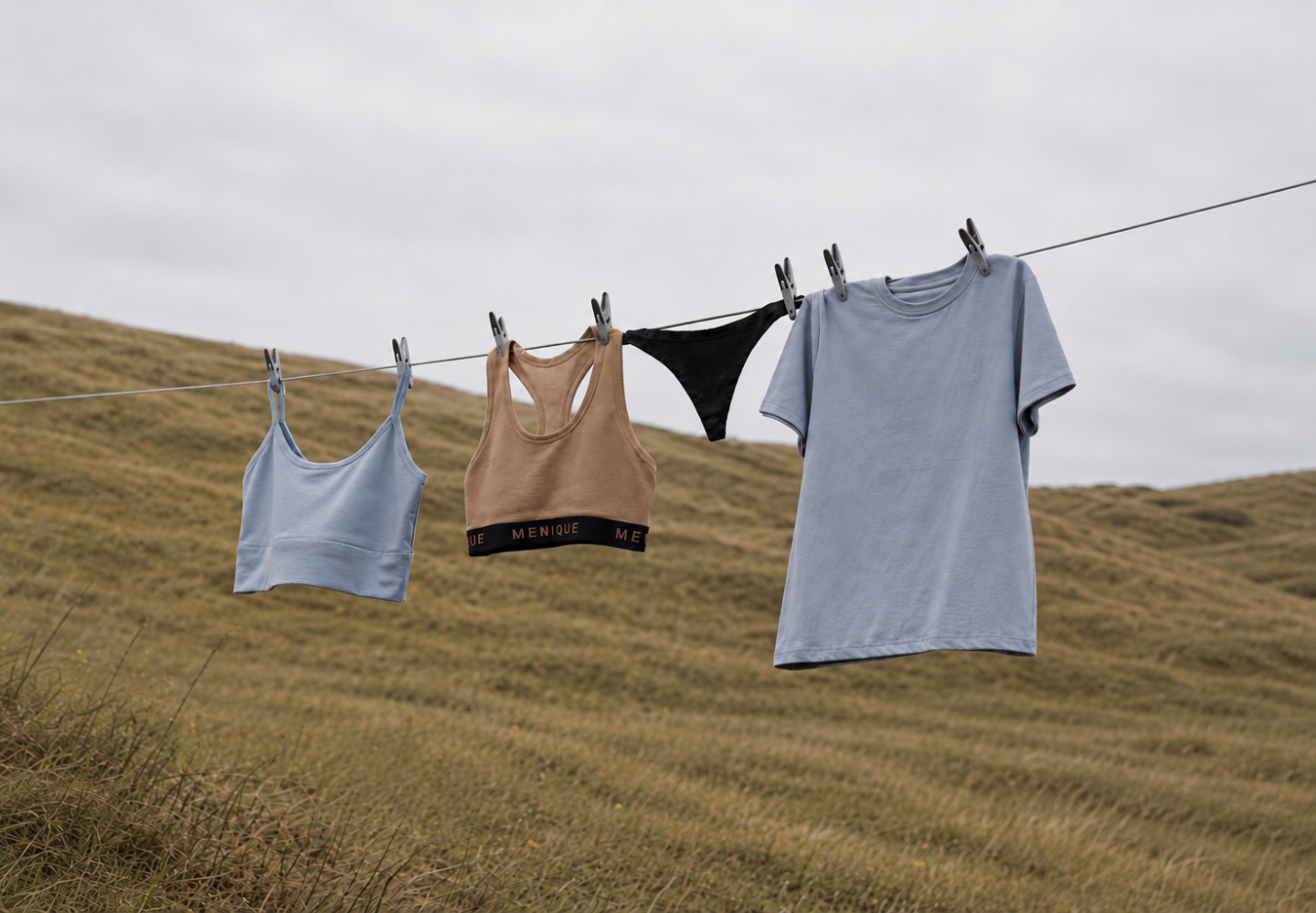 Four pieces of clothing, including a light blue t-shirt and a tan sports bra, hang from a clothesline secured by gray pegs. The garments dry outdoors against a backdrop of rolling grassy hills under an overcast sky.