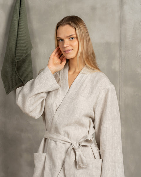 A woman with blonde hair wearing a natural color 100% linen menique robe with a tied belt, touching her face, against a textured grey background.