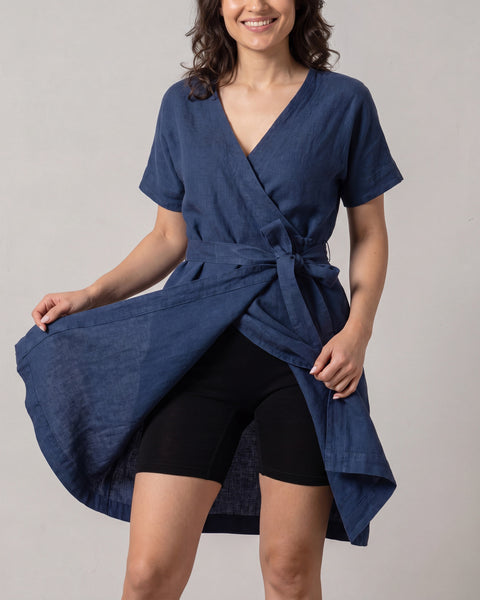 A smiling woman wearing a short-sleeved, navy blue linen wrap dress holds the front of her skirt open to reveal fitted, mid-thigh black slip shorts worn underneath.