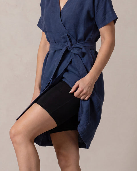 A mid-body shot of a woman wearing a blue dress, lifting the side of her skirt while raising her bent right knee to display the coverage and fit of the black slip shorts beneath her dress.