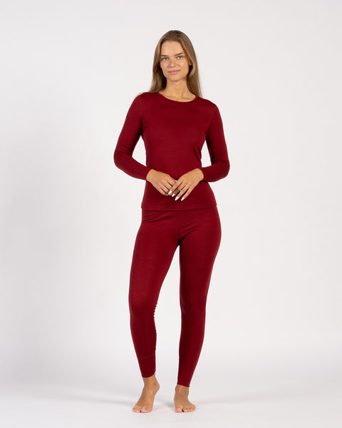 A studio portrait of a barefoot blonde woman standing and looking directly at the camera. She is wearing a matching royal cherry deep red long-sleeve thermal crew neck top and leggings, with her hands gently clasped in front of her waist against a plain light grey background.