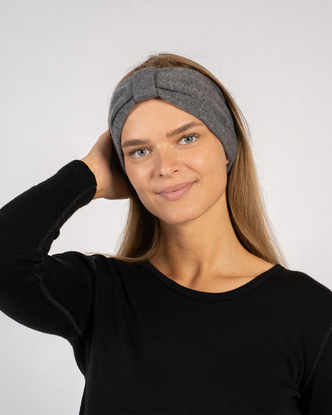 Woman wearing a dark grey headband with a front knot detail, dressed in a black long-sleeve top, smiling softly while touching her hair against a plain light background.