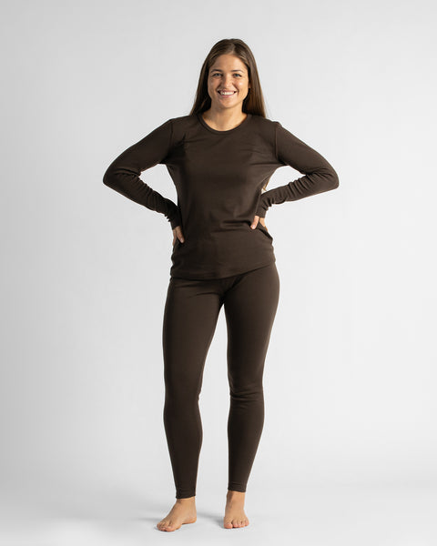 A full-length portrait of a barefoot woman standing against a white studio background with hands on her hips, wearing a fitted dark brown thermal loungewear set.
