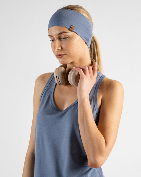 A studio portrait of a blonde woman looking down and to the left against a plain light grey background. She is wearing a dusty blue v-neck sleeveless sport top and a matching wide dusty blue headband, with one hand resting on the beige headphones around her neck.