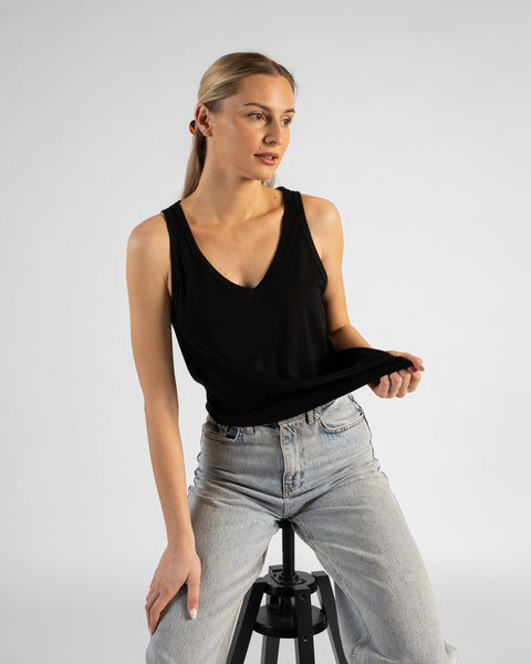 A studio portrait of a blonde woman sitting on a black stool, looking off to her right. She is wearing a black sleeveless V-neck top and light wash denim jeans, gently pulling the hem of her top with one hand while her other hand rests on her leg against a plain light grey background.