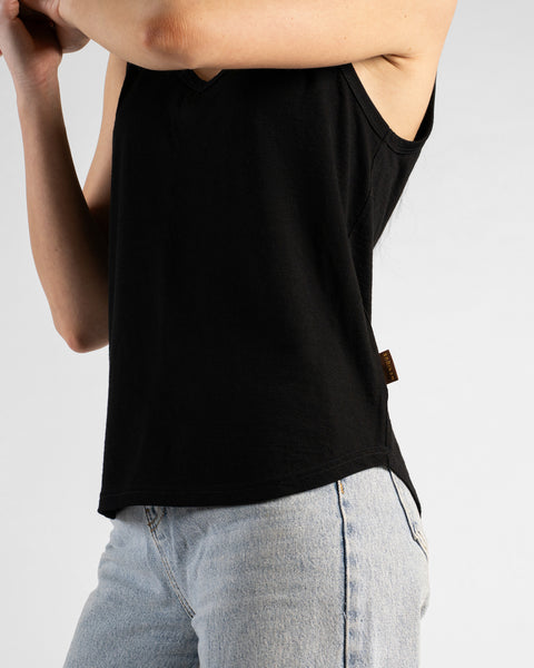 A close-up side profile shot focusing on the torso of a woman against a plain light grey background. She is wearing a black sleeveless V-neck top featuring a small brown logo tag on the side seam, paired with light wash denim jeans, with her arms crossed lightly in front of her.