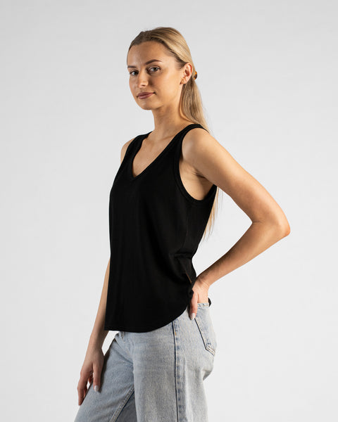 A studio portrait of a blonde woman standing in a three-quarter profile and looking directly at the camera. She is wearing a black sleeveless V-neck top and light wash denim jeans, with one hand resting on her hip against a plain light grey background.