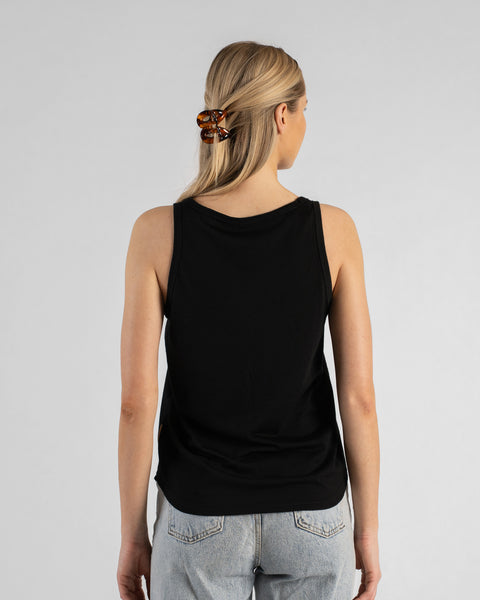 A back view studio shot of a blonde woman standing against a plain light grey background. She is wearing a black sleeveless top and light wash denim jeans, with her hair styled half-up using a brown tortoiseshell claw clip.