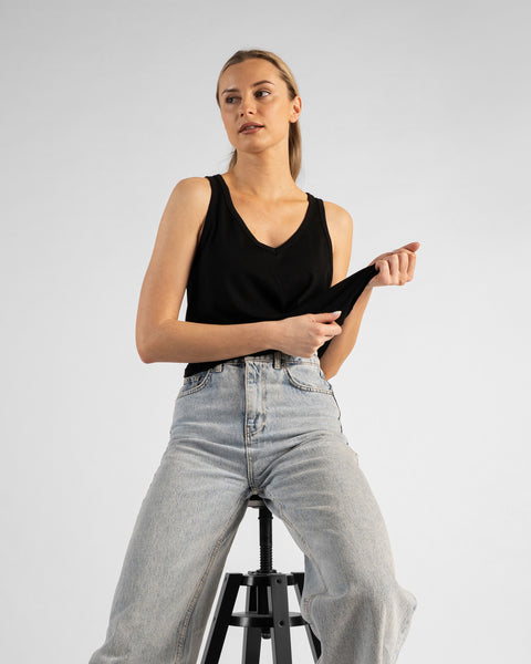 A studio portrait of a blonde woman sitting on a black stool and looking off to her right. She is wearing a black sleeveless V-neck top and light wash denim jeans, lightly holding the hem of her top with both hands against a plain light grey background.