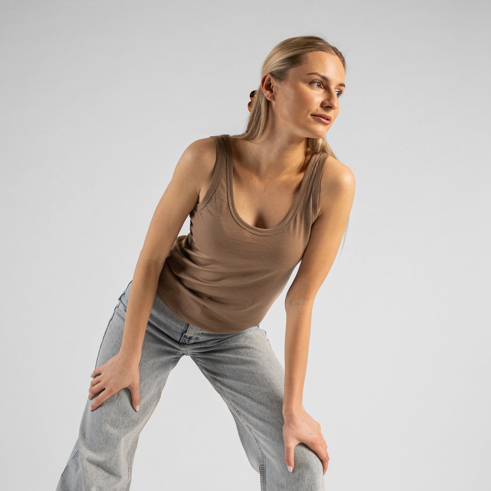 A studio portrait of a blonde woman standing and looking directly at the camera with a subtle smile. She is wearing a beige sleeveless scoop-neck top and light wash denim jeans, with one hand casually resting in her front pocket against a plain light grey background.
