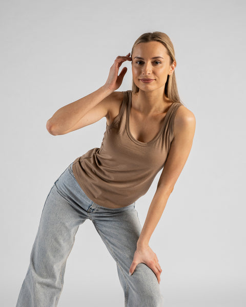 A studio portrait of a blonde woman leaning slightly forward and looking directly at the camera. She is wearing a beige sleeveless scoop-neck top and light wash denim jeans, with one hand resting near her temple and the other on her thigh against a plain light grey background.