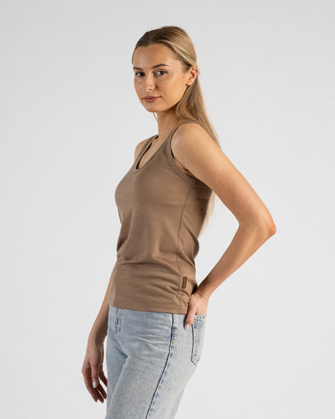A studio portrait of a blonde woman standing in a three-quarter profile and looking directly at the camera. She is wearing a beige sleeveless scoop-neck top and light wash denim jeans, with one hand resting in her front pocket against a plain light grey background.