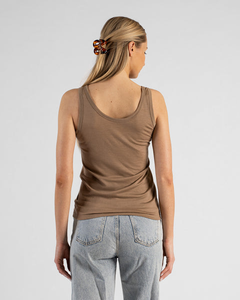 A back view studio shot of a blonde woman standing against a plain light grey background. She is wearing a beige sleeveless top and light wash denim jeans, with her hair styled half-up using a brown tortoiseshell claw clip.