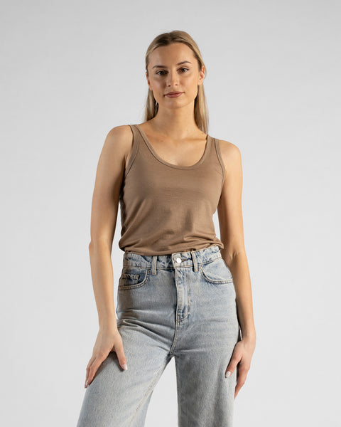 A studio portrait of a blonde woman standing and looking directly at the camera with a subtle smile. She is wearing a beige sleeveless scoop-neck top and light wash denim jeans, with one hand casually resting in her front pocket against a plain light grey background.