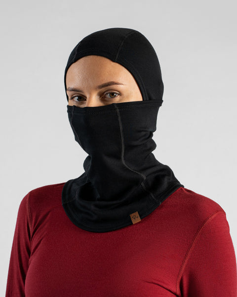 A studio portrait of a blonde woman looking at the camera, wearing a burgundy long-sleeved shirt and a black balaclava fully covering her head and lower face, leaving only her eyes exposed against a light grey background.