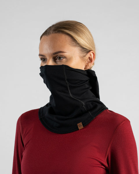 A studio portrait of a blonde woman looking to the left, wearing a burgundy long-sleeved shirt and a black balaclava pulled up to cover her nose and mouth, with the hood section pushed back off her head.