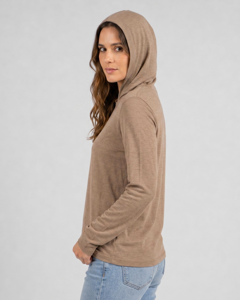 A side-profile studio portrait of a woman looking back over her shoulder at the camera against a plain light grey background. She is wearing a lightweight beige long-sleeved hoodie with the hood pulled up over her head and light wash denim jeans.