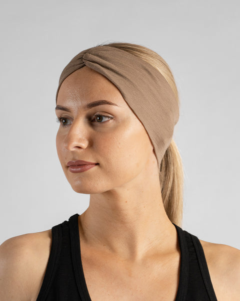 A studio portrait of a blonde woman looking to her right against a plain light grey background. She is wearing a black sleeveless top and a beige merino ribbon headband with the twisted ribbon detail centered on her forehead.