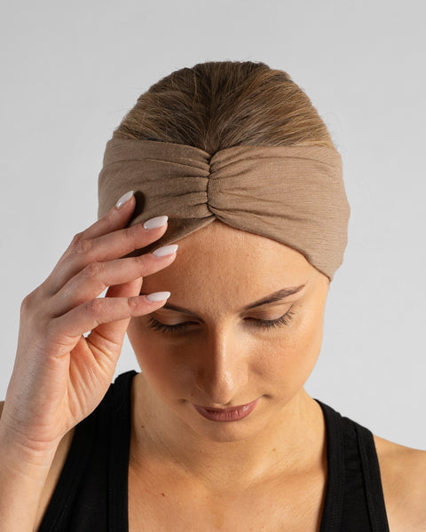 A studio portrait of a blonde woman looking downward with her right hand gently resting on her forehead. She is wearing a black sleeveless top and a beige merino ribbon headband with the twisted ribbon detail centered on her forehead, set against a plain light grey background.