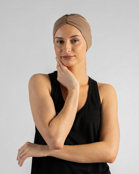 A studio portrait of a blonde woman looking directly at the camera with a subtle smile. Her arms are crossed, and her right hand gently touches her jawline. She is wearing a black sleeveless top and a beige merino ribbon headband against a plain light grey background.